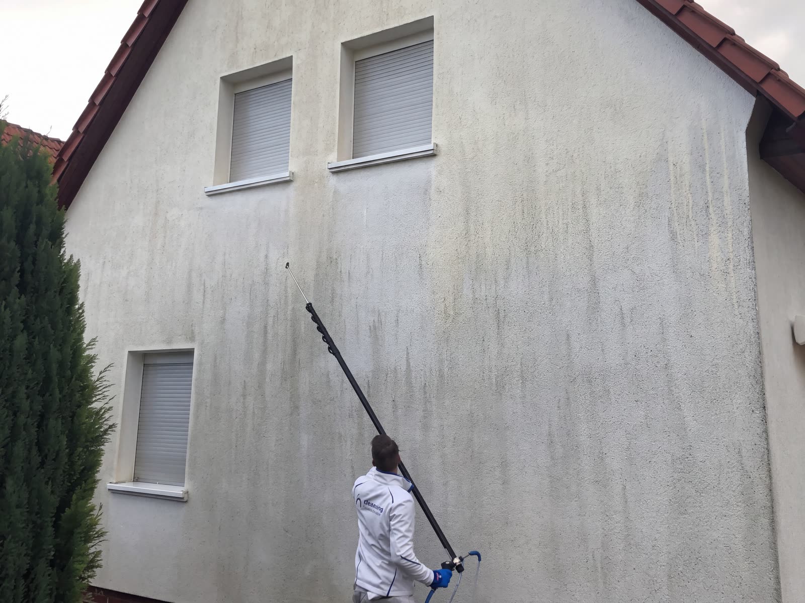 Mitarbeiter trägt das Reinigungsmittel mit der Teleskoplanze vom Boden aus an einer hohen Putzfassade auf.