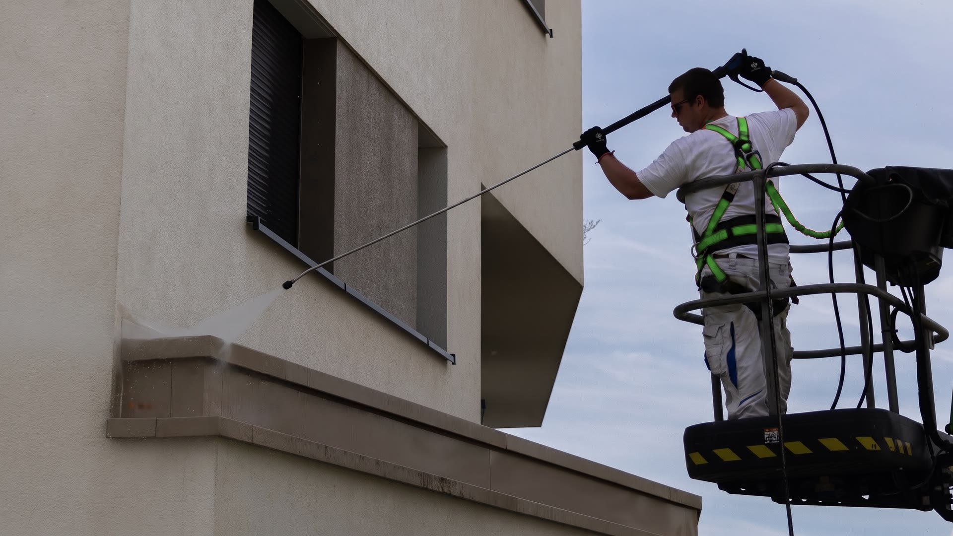 Ein Mitarbeiter reinigt vom Korb einer Arbeitsbühne aus die obere Fassade eines Mehrfamilienhauses; unter dem Wasserstrahl wird der Putz sichtbar heller.